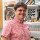 Head and upper body of a white man with short dark hair and glasses wearing a pink short sleeved shirt and smiling. Behind him are bookshelves. 