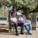 Three students chat and sit on benches in a shady spot near the UC Davis Quad. A “No Bicycle Parking” sign is visible nearby, along with tall trees and a white tent in the background.