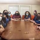 A group of masked UC Davis student staff and leaders sit around a large conference table inside the Native Nest. They smile behind their masks, dressed in matching navy UC Davis polos and casual clothes.