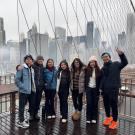 A group of seven young adults stands close together, smiling on the Brooklyn Bridge in New York City. The overcast skyline and tall buildings are visible behind them through the bridge’s cables. Everyone wears winter jackets, hats, and scarves. The bridge's wooden walkway appears wet and one person on the far right flashes a peace sign.