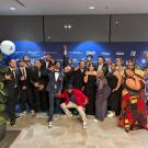 A large group of students and staff pose in formal wear at a blue-carpet event. They stand in front of a branded backdrop with sponsor logos. Many are making fun or celebratory gestures and smiling. Blue and white balloon arrangements frame the scene.