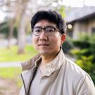 A man with glasses stands outdoors, wearing a light jacket, with greenery in the background.