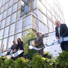 Outdoor setting, large glass building in background. Four people stand in a row cutting a ribbon into pieces. 