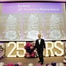 A man in a tuxedo stands on a stage festooned with grape-like orbs, a lit sign reading "25 YRS" and a banner reading, "Big Bang! 25th Anniversary Physical Awards"