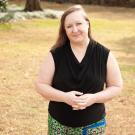 A woman with shoulder length blonde hair wearing a black sleeveless top and patterned skirt stands outdoors with her hands clasped in front. 