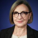Woman with short gray hair and glasses, wearing a black blazer and colorful necklace, smiling.