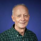 Smiling older man with gray hair, wearing a green checkered shirt, against a blue background.