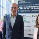 Left, a man with cropped hair and glasses wearing a grey jacket; right, a shorter woman with shoulder length brown hair, glasses and a pink jacket. They stand in front of a poster for the 25th Anniversary of the Bryan Miller Symposium with a list of sponsors. 