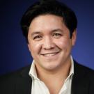 Smiling man with dark hair, wearing a black blazer over a white shirt, against a blue background.