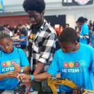 Two children in light blue UCSTEM shirts at a table with machinery. An adult in a check shirt stands behind them pointing to details. 