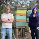 Sascha Nicklish in ponytail, white t-shirt and jeans stands with Julia Fine in black shirt and pants and purple hair stand on either side of a wooden, mint green and yellow bee hive. 