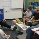 Seven students sit in a circle, laugh and eat snacks together in a UC Davis lounge. 