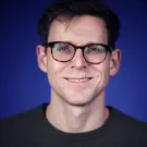 A smiling person with glasses against a blue background.