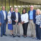 A group of six professionally dressed stand outside a venue, smiling and staring at the camera. The group includes 