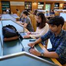 Two students one in a long sleeve white top and one in a blue and yellow plaid shirt, study on their computers in Shields Library Main Reading Room.