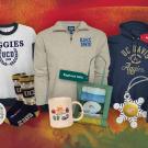A collection of UC Davis merchandise displayed on a red, orange, and green textured background. The items include a white sweatshirt with navy trim featuring 'AGGIES UCD 1908' with a laurel wreath design, a beige quarter-zip pullover with UC Davis logo, and a navy blue hoodie with gold 'UC DAVIS Aggies' lettering. Accessories include UC Davis patterned socks, a holiday ornament with a Nordic-style pattern, a ceramic mug with geological illustrations, a green tote bag featuring an Egghead sculpture design, a