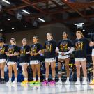 Inside a basketball gymnasium, a row of women basketball players face the camera. They're all wearing t-shirts that read 'Black Futures Month'.