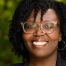 A woman wearing a black blazer smiles in front of a background of greenery