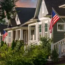 Suburban street scene with modest Craftsman style homes, American flags and green lawns.