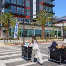Two people push moving carts across a street toward housing at Aggie Square