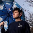 Colin McCarthy stands outdoors on a rainy day, holding an umbrella printed with blue sky and clouds. Bare trees are visible in the background, and there are blurry raindrops on the camera lens.