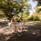 Two students bike along a campus road against a blurred background of trees
