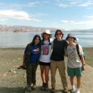 Four smiling people stand side by side on a lakeshore with their arms around each other. They are dressed in casual outdoor clothing and hats, with a scenic lake and mountain range in the background under a blue sky with scattered clouds.