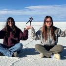 "On a sunlit salt flat, two women wearing sunglasses sit on the ground, playfully holding up smaller figures in their hands through forced perspective. The smaller figures in the background stand in yoga poses, with the salt flats stretching out beneath a wide, clear sky."