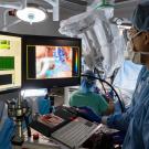Surgeon look at computer screens and uses mouse inside operating room. A large surgical robot is seen in the background, alongside another surgeon.
