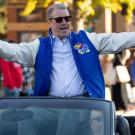 A man in a University of Kansas jacket stands up in the back of a convertible with his arms held out wide, in a parade.