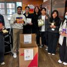 A group of seven people holding food and hygenic products near a donation box.