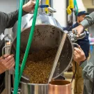 Two men pour grain from a large container into a smaller pot in a brewing facility.