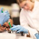 A student in a white lab coat, blue gloves and safety goggles carefully uses a pipette while another student in a lab coat observes in the background in a laboratory setting.