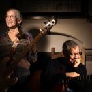 A photograph features two musicians in a warmly lit room with a fireplace in the background. The woman on the left, wearing glasses and a colorful patterned blouse, is holding a bass guitar with a leather strap, smiling brightly while looking off to the side. Her curly gray hair is pulled back. The man on the right, wearing glasses and a black sweater, sits on a wooden chair with his elbow resting on the guitar he is holding. He appears thoughtful, with a gentle smile and his hand resting on his cheek.