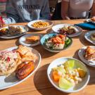 Several different plates of food sit atop an outdoor picnic table bathed in sunlight, with patrons on sitting on either side of the table. 