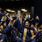 Graduates in caps and gowns celebrate joyfully, waving their hands in excitement.