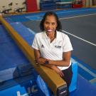 Female in white Aggie Athletics polo shirt, one arm atop balance beam in gym