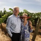 Portrait of a couple standing in a vineyard on a sunny day. The man wears a light colored shirt and glasses. The woman wears a sundress and large-brimmed straw hat.