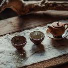 Ceramic teapot with two small cups on a textured wooden surface, framed by a branch.
