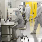 Researchers wearing full cleanroom suits work inside a bright, sterile laboratory. One person sits at a metal workstation using a computer, while others walk or operate equipment in the background. Rows of storage lockers line one side of the room, and large yellow doors are visible at the back.