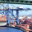 Photo of a large container ship, seen from the stern, and docked next to a large crane. Many other containers are stacked high on the dock, and the lower trusses of a green bridge can be seen spanning over the ship and port.