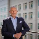 A tieless man wearing a suit stands on a balcony, juxtaposed against a building facade