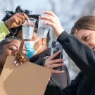 Three students closely examine clear plastic cups filled with water, holding them up to the light. One student points at a cup while holding a clipboard, another wears a face mask in the background, and a third uses a smartphone to take a photo or video. The scene takes place outdoors on a sunny day.