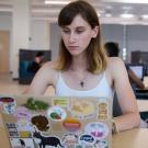 Undergraduate Laila Penny sits at a table in a bright, modern study space, focused on her laptop. She has light skin, straight brown hair with blonde tips, and wears a white tank top and a lightning bolt necklace. Her laptop is covered with colorful stickers, including animals, food, and slogans like “Black Lives Matter.” Other students are visible in the background, working or socializing in small groups. 