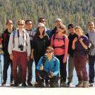 Group of hikers posing together by a lake, surrounded by trees and mountains.