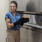 woman in blue shirt and tan pants smiles while holding loaf of freshly baked bread pulled from large metal oven