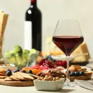 A glass of red wine sits on a table with a spread of fruit, cheeses, nuts and crackers.