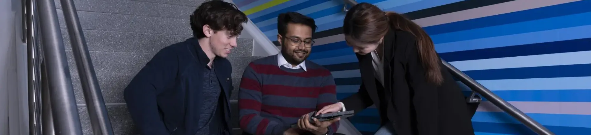 three students sitting on steps talking and looking at a device