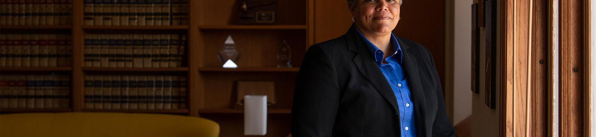 Judge Kelli Evans, wearing a bright blue button-up and black jacket, stands in the UC Davis law school building for a photo. Light flows in through a window, and a mustard couch and shelves of old law books are in the background.