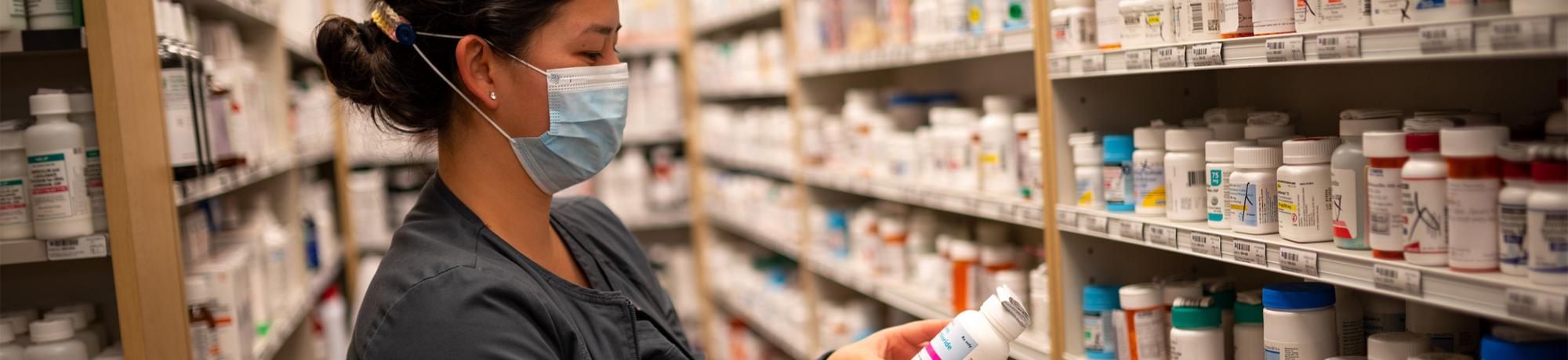 A pharmacist in a blue face mask stands between rows of shelving holding prescription medication and examines one of the bottles.